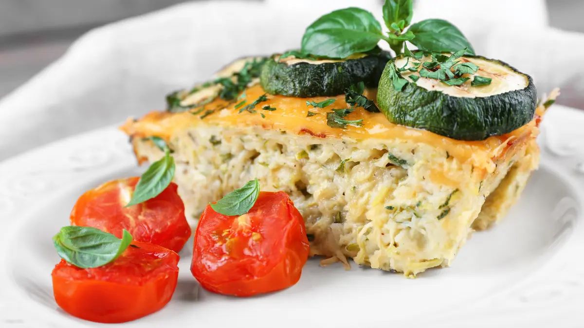 Ein Stück Gemüsetarte auf einem Teller, garniert mit Zucchini, Tomate und Basilikum