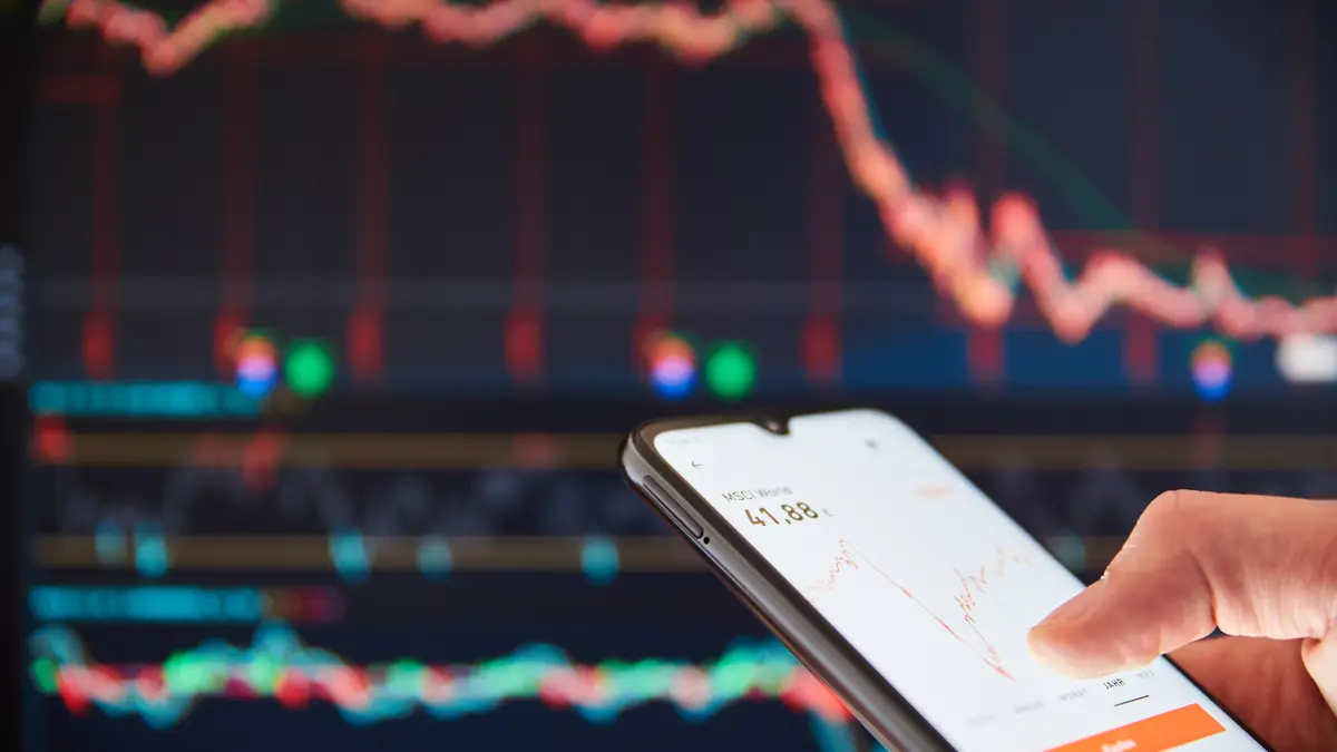 Nahaufnahme eines Smartphones, das in einer Hand gehalten wird und einen roten, absteigenden Graphen auf dem Bildschirm anzeigt. Im Hintergrund ist ein größerer Bildschirm mit einem ähnlichen Graphen zu sehen, der ebenfalls einen Abwärtstrend zeigt.