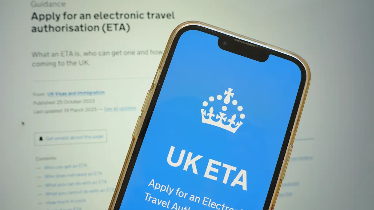 Ein Smartphone zeigt die UK-Regierungswebsite zur Beantragung der elektronischen Reisegenehmigung (ETA) für das Vereinigte Königreich an. Im Hintergrund ist ein verschwommener Bildschirm mit weiteren Informationen zur ETA sichtbar.