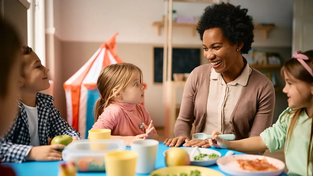 Eine Erzieherin sitzt mit Kindern im Kindergartenalter am Esstisch