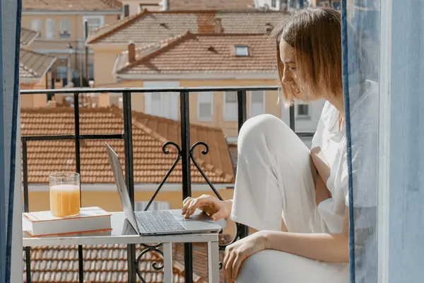 Frau sitzt mit Laptop an einem Tisch auf einem Balkon