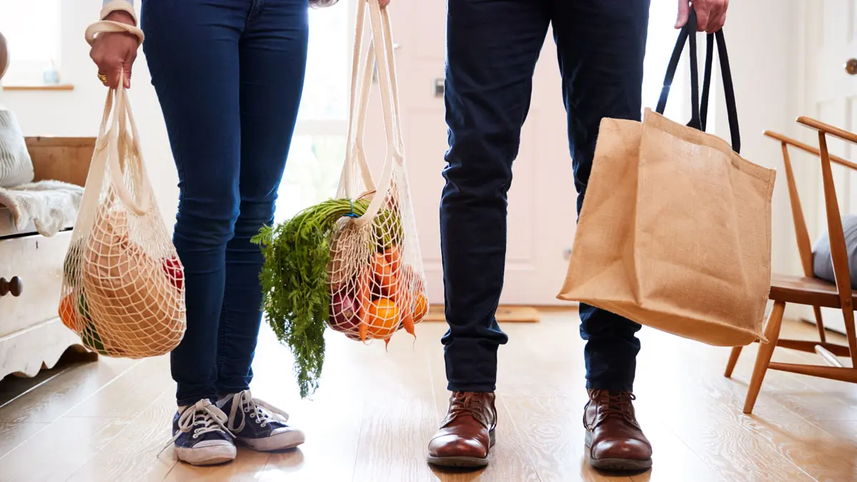 Zwei Personen mit Lebensmitteln in Einkaufsnetzen und -taschen stehen in einer Küche, man sieht nur ihre Beine