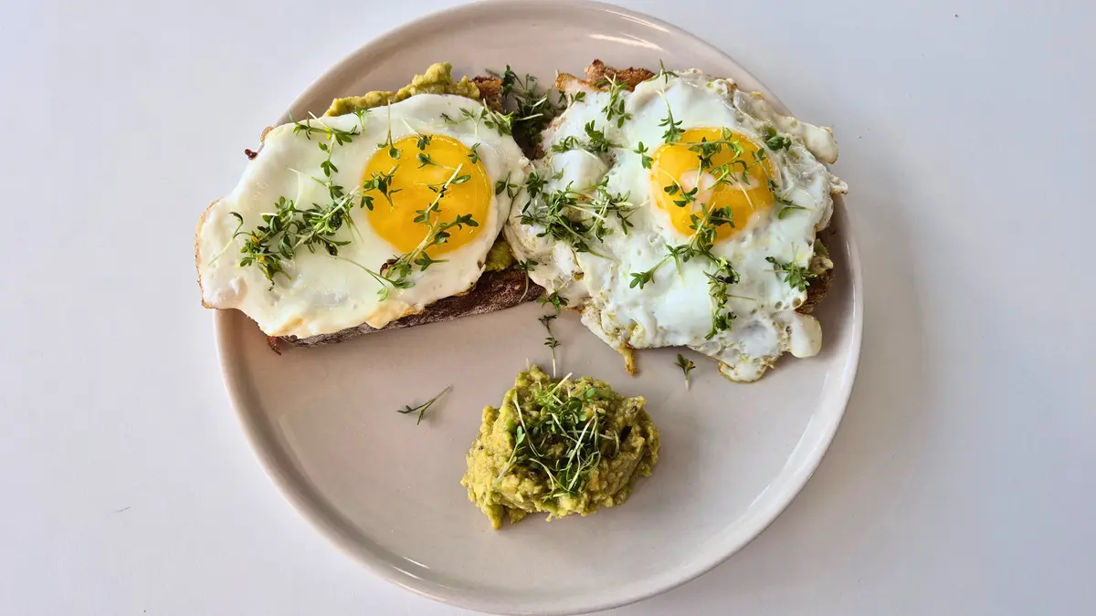 Zwei belegte Brotscheiben auf einem Teller: zwei Spiegeleier mit Kresse und eine kleine Portion Avocado-Aufstrich.