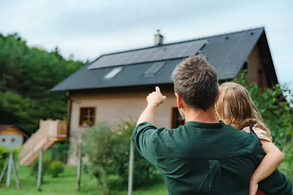 Vater mit Kind auf dem Arm zeigt auf Haus mit Photovoltaik Dach