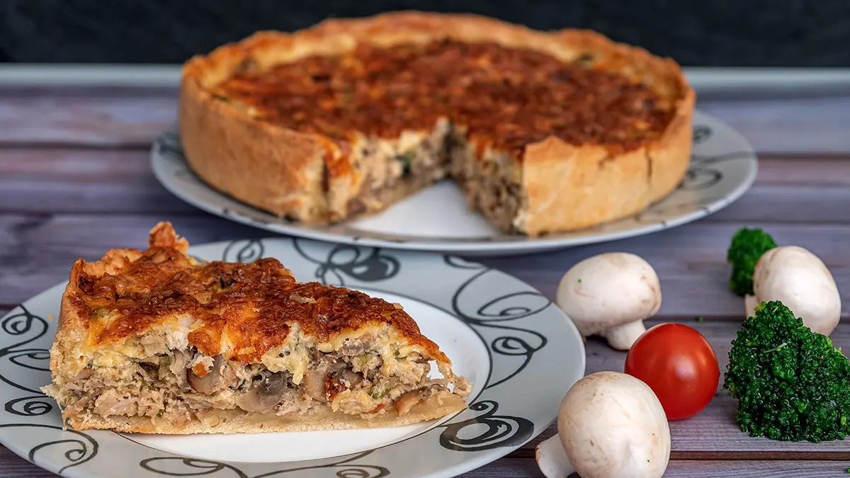 Eine Quiche auf einem Teller, ein Stück der Quiche auf einem weiteren Teller, daneben liegen einige Chamignons, Tomaten und Brokkoli.