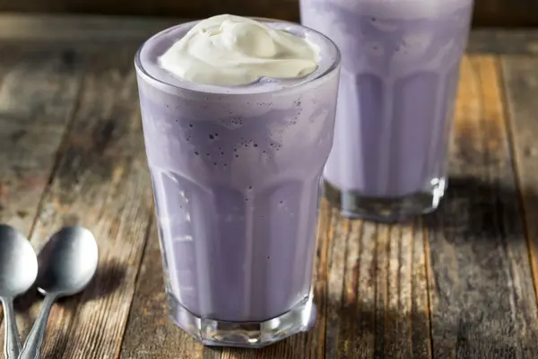 Zwei mit der lilafarbenen Yams-Wurzel Ube gefärbte Milchshakes mit Sahne stehen auf einem Holztisch neben zwei Löffeln