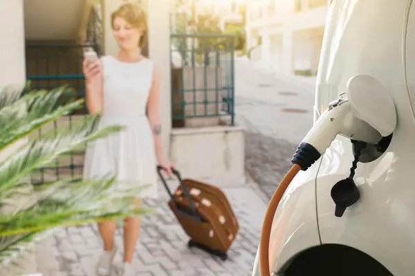 Frau mit Handy in der Hand und weißem Kleid läuft auf ein weisses E-Auto zu, welches an einer Ladesäule geladen wird.
