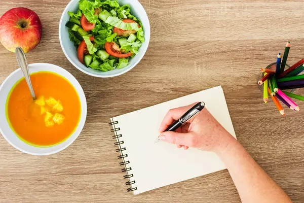 Ein Tisch mit Suppe und Salat in Schälchen, einem Apfel und Buntstiften. Eine Hand schreibt auf einen Block
