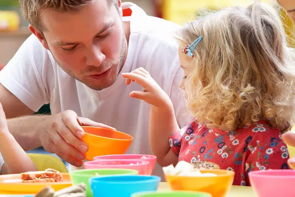 Ein Mann sitzt mit Kindern an einem Kindertisch mit bunten Schüsseln beim Essen
