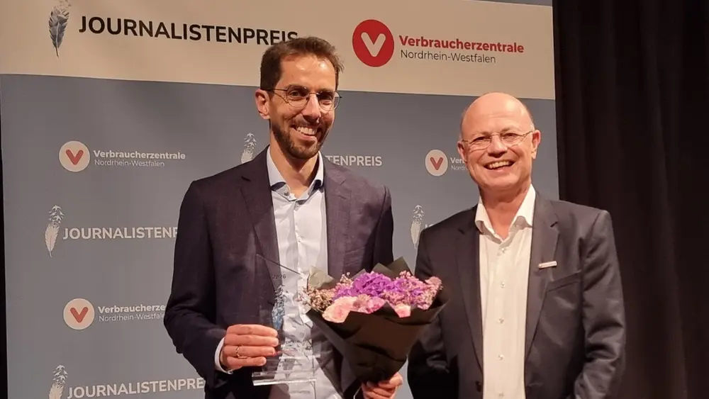 Zwei Männer blicken lächelnd in die Kamera, einer von ihnen hält einen Blumenstrauß und eine Trophäe in den Händen.