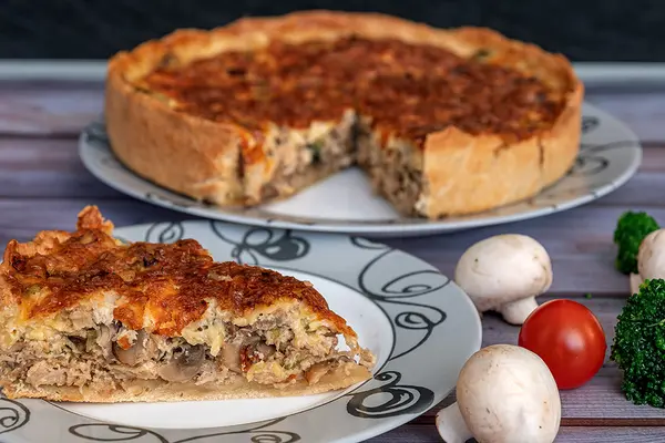 Eine Quiche auf einem Teller, ein Stück der Quiche auf einem weiteren Teller, daneben liegen einige Chamignons, Tomaten und Brokkoli.