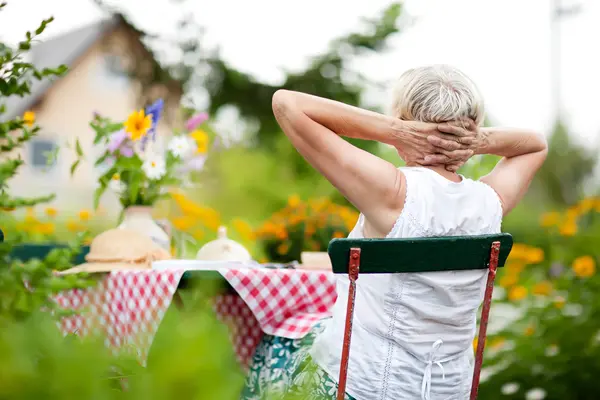 Seniorin sitzt im Garten und schaut Haus an