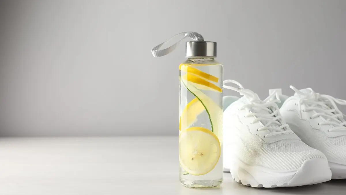 Eine Trinkflasche, die mit Wasser und Zitronenscheiben gefüllt ist, steht neben einem Paar weißer Turnschuhe.