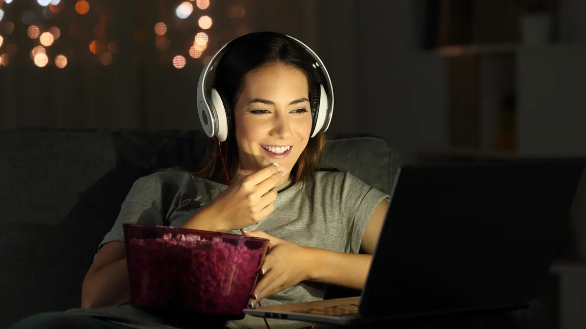 Eine lächelnde Frau mit weißen Kopfhörern und einer Schale Popcorn , die nachts im Internet Fernsehen schaut.