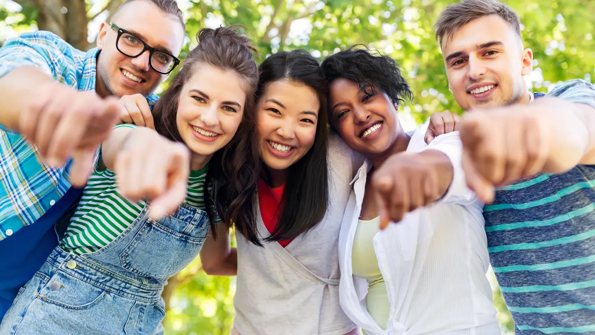 Grupp aus 5 jungen Menschen schaut in die Kamera und zeigt mit Fingern auf Kamera.