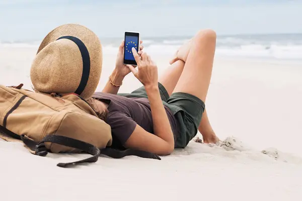 Eine Frau liegt im weißen Sand eines Strandes, Wellen unscharf im Hintergrund. In der Hand hält sie ein Smartphone mit der EU-Flagge auf dem Display.