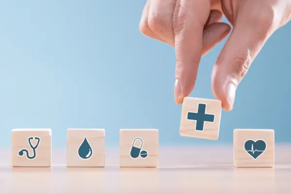 eine Hand nimmt einen Würfel mit einem blauen Kreuz aus einer Reihe von Würfeln mit verschiedenen Symbolen aus dem Gesundheitswesen. Eine Hand nimmt einen Würfel mit einem Plus.
