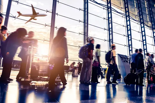 in geschäftiges Flughafenterminal mit Passagieren in Bewegung, die als verschwommene Silhouetten vor einer großen Glasfassade dargestellt sind. Im Hintergrund ist ein Flugzeug beim Start durch das Fenster zu sehen, während helles Sonnenlicht den Raum flutet und lange Schatten sowie Lichtreflexe auf dem glänzenden Boden erzeugt.