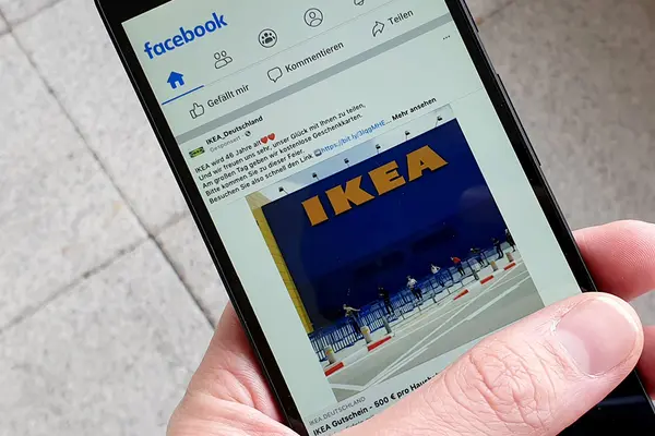 Hand hält Smartphone, Display zeigt einen Beitrag mit dem Foto eines Ikea-Gebäudes auf Facebook
