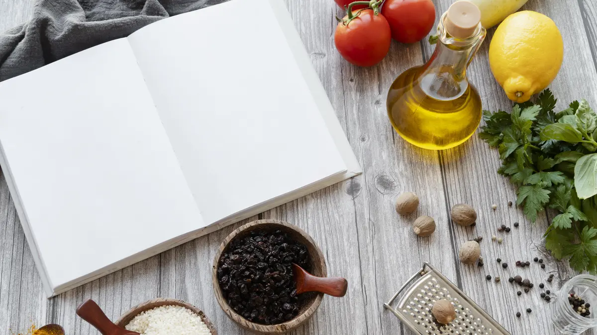 Ein aufgeschlagenes leeres Rezeptbuch umrandet von Lebensmitteln (Teelöffel aus Holz mit Kurkuma, kleines Holzschälchen mit trockenem Reis, kleines Holzschälchen mit Rosinen, Tomaten, Zitrone, Petersilie, Glasfalsche mit Öl, Muskatreibe und Muskatnüssen, Pfefferkörner)