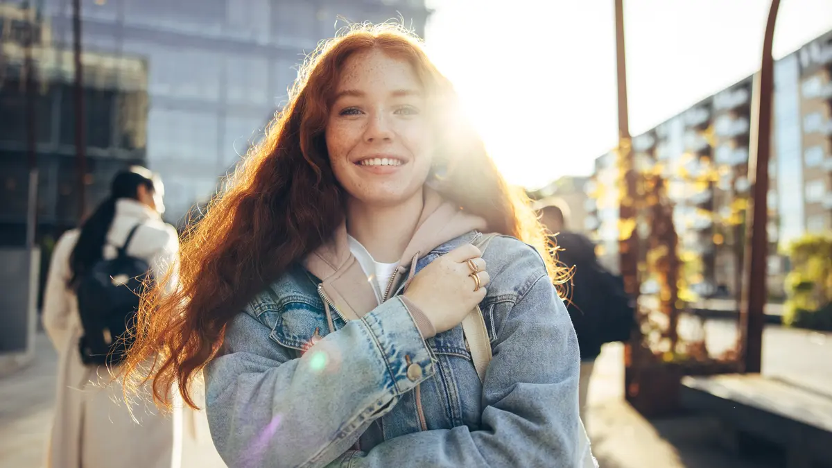 Eine junge rothaarige Frau lacht in die Kamera. Hinter ihr ist ein verglastes Gebäude und die Sonne scheint. Sie trägt einen Rucksack und im Hintergrund sieht man weitere Personen mit Rucksack.