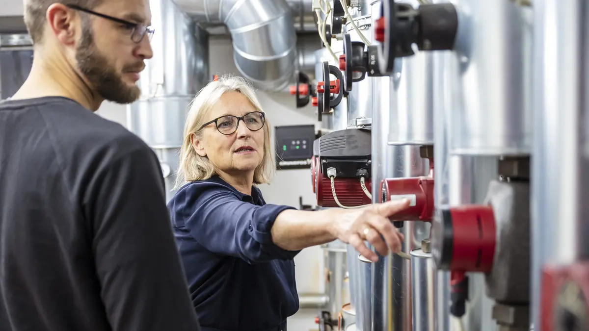 Eine Energieberaterin erläutert einem Verbraucher eine technische Anlage im Keller.