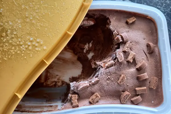 Eine geöffnete Schokoladeneispackung steht auf der Küchenarbeitsplatte. Auf der Deckelinnenseite und auf der Oberfläche des Speiseeises sind Eiskristalle zu sehen.