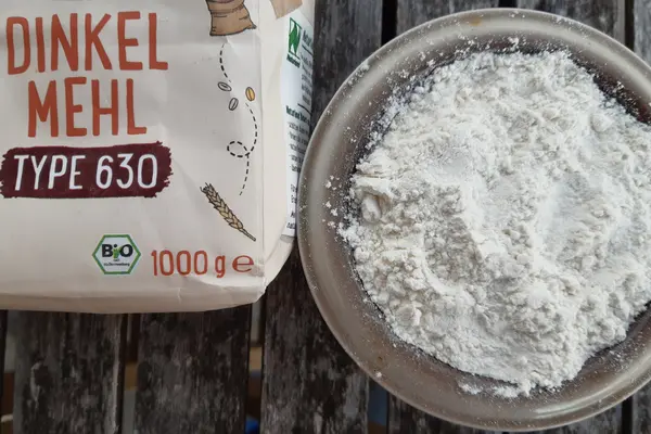 Eine Tüte mit Dinkelmehl liegt neben einer Glasschüssel, in der Dinkemehl enthalten ist.