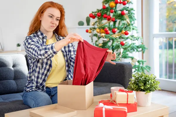 Frau sitzt vor einem Weihnachtsbaum auf der Couch und öffnet ein Geschenk, das ihr nicht gefällt