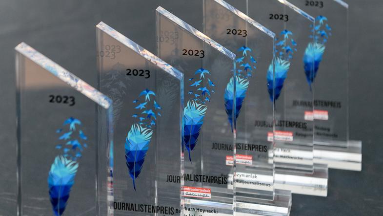 Sechs Trophäen aus Acrylglas mit der Zahl 2023 über einer blauen Feder, Namen der Preisträger:innen und Logo der Verbraucherzentale Nordrhein-Westfalen