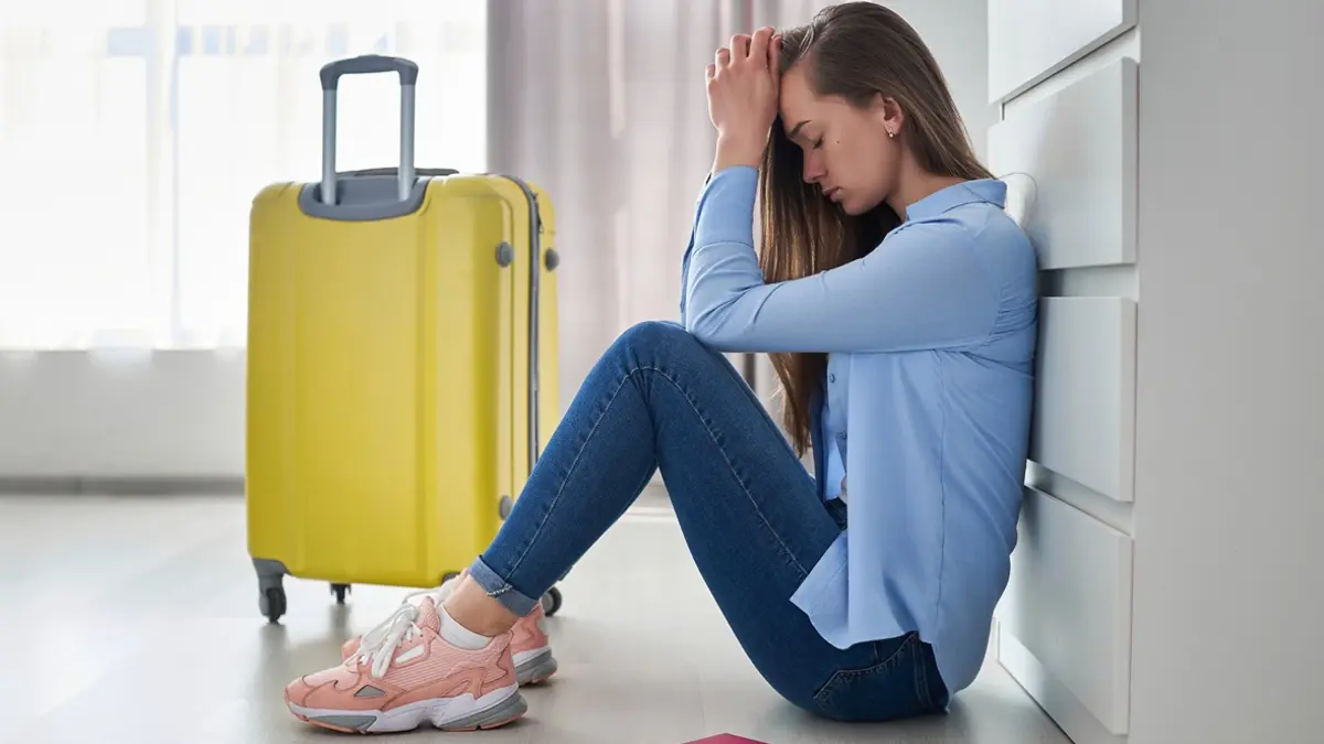Eine junge Frau sitzt mit gesenktem Kopf und den Händen an der Stirn neben einem gelben Rollkoffer auf dem Boden; vor ihr liegt eine rote Reisepasshülle.