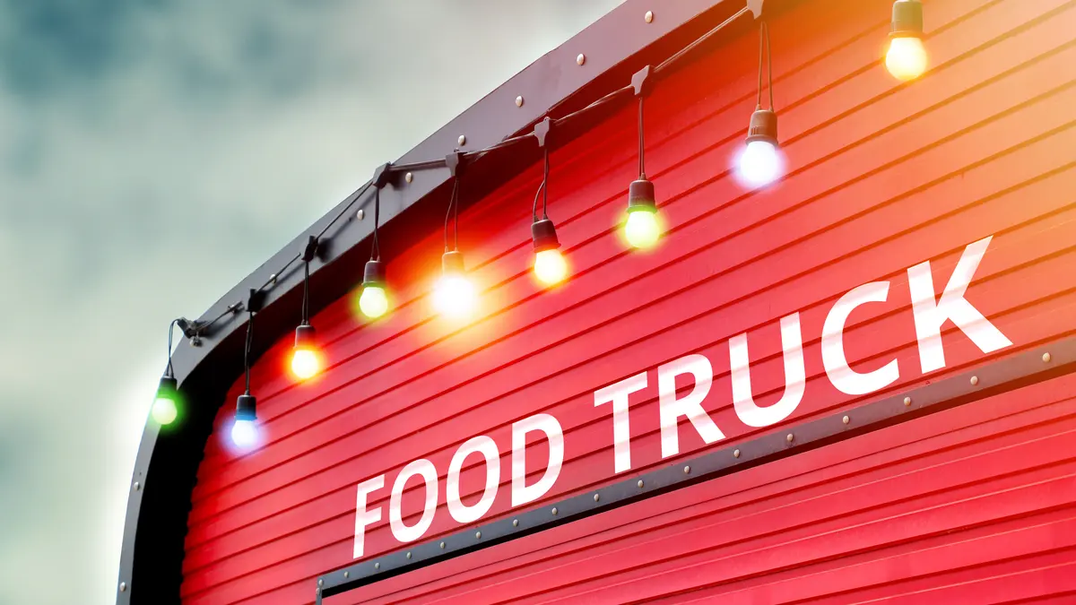 Rote Seitenwand eines Food Trucks mit der weißen Aufschrift "FOOD TRUCK", darüber eine Lichterkette mit weißen Glühbirnen vor bewölktem Himmel.