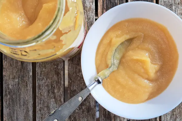 Ein geöffnetes Glas Apfelmus steht neben einer weißen Schüssel mit Apfelmus