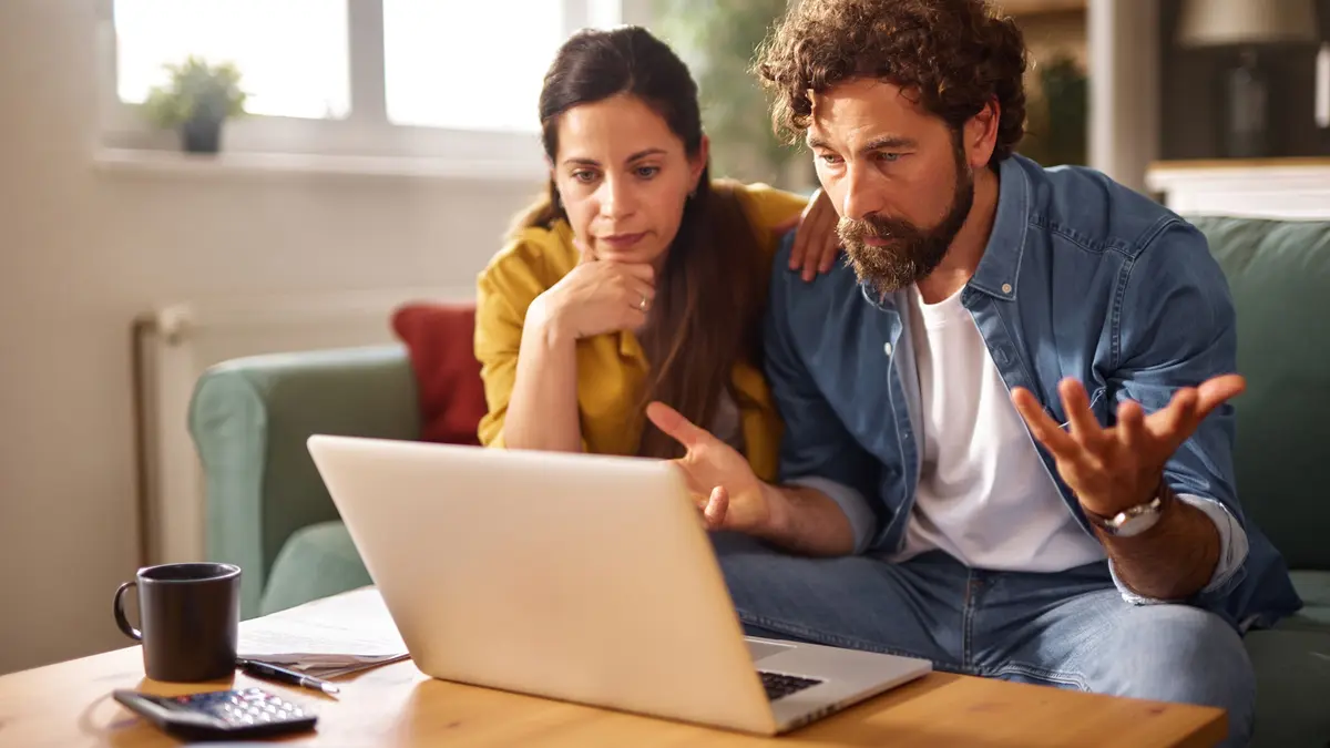 Ein Paar wirkt gestresst, während es auf einer Couch sitzt und auf einen Laptop schaut. Sie sind von Dokumenten und einer Kaffeetasse umgeben, was auf eine ernste Diskussion über finanzielle Probleme hindeutet.