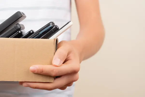 Nahaufnahme einer Person, die einen kleinen Pappkarton hält, der mit mehreren alten Mobiltelefonen und Smartphones gefüllt ist.