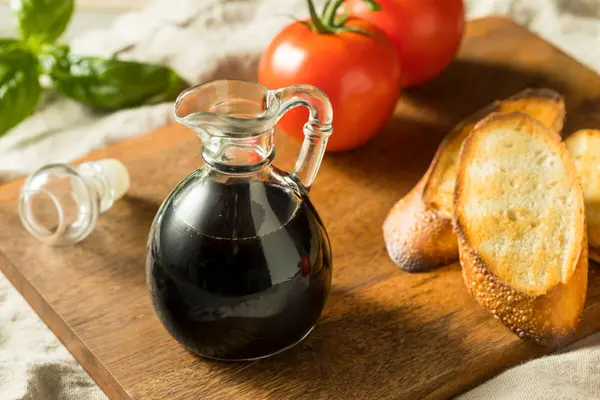Ein Glaskännchen mit Balsamico Essig steht auf einem Holzbrett, daneben liegen zwei Scheiben getoastetes Baguette und zwei Tomaten
