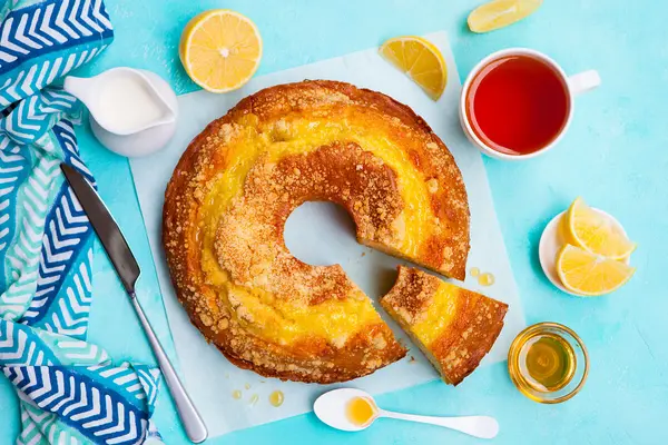 Ein Rührkuchen von oben, daneben befinden sich eine aufgeschnittene Zitrone, eine Tasse Tee, ein Kännchen Milch, ein Messer und ein Löffel.