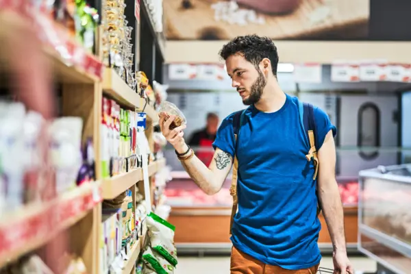 Ein junger Mann steht vor einem Regal im Supermarkt, hat ein Produkt in der Hand und liest von der Verpackung 