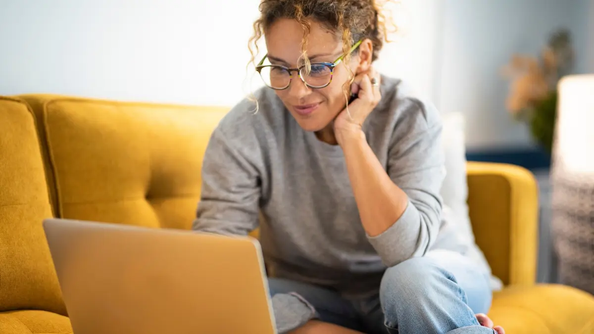 Frau mit Brille sitzt auf einem gelben Sofa und arbeitet mit einem Laptop.
