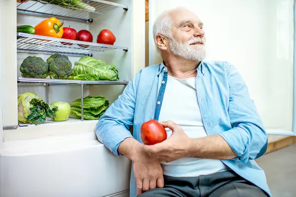 Senior sitzt am offenen Kühlschrank mit Obst und Gemüse