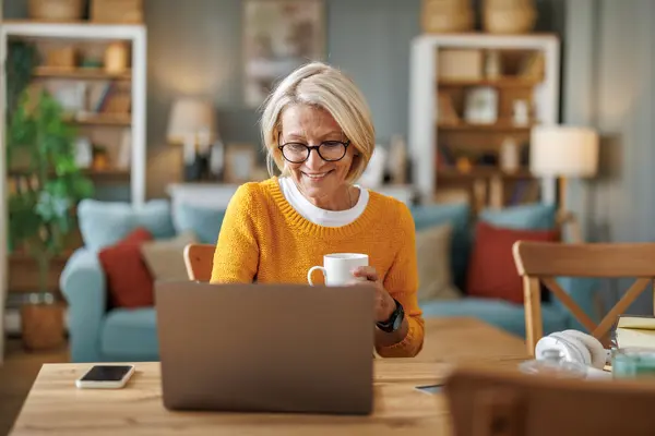 Eine blonde Frau mittleren Alters sitzt am Esstisch vor ihrem Laptop im Wohnzimmer. Sie hält eine weiße Tasse in ihrer Hand, schaut auf das Display und lächelt.