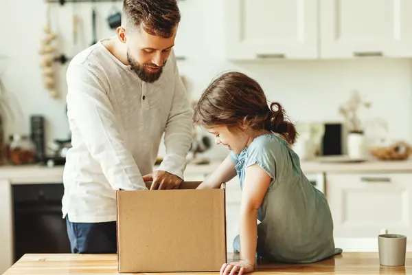 Ein Mann und Mädchen sind in der Küche, der Mann steht hinter der Küchentheke und das Mädchen sitzt darauf. Neben ihr steht ein Paket, das Vater und Tochter gemeinsam auspacken.