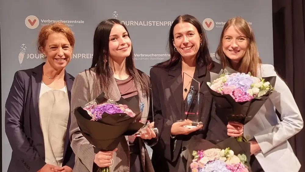Vier Frauen stehen nebeneinander und lächeln in die Kamera. Drei von ihnen halten Blumensträuße und Trophäen in den Händen.