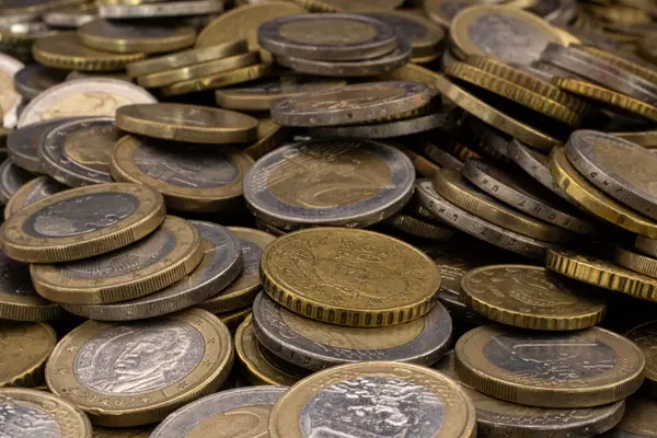 Eine Nahaufnahme zeigt einen großen, ungeordneten Haufen verschiedener Euro-Münzen, die übereinandergestapelt sind. Im Vordergrund sind deutlich 1-Euro- und 2-Euro-Münzen sowie goldfarbene Cent-Stücke (wahrscheinlich 50 Cent) zu erkennen. Die Münzen weisen Gebrauchsspuren und eine matte Textur auf, wobei das Licht die geprägten Ränder und Oberflächendetails hervorhebt.