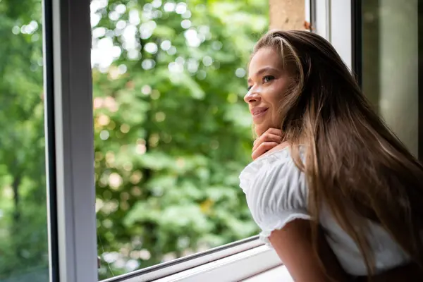 Eine junge Frau lehnt sich auf die Fensterbank eines offenen Fensters und blickt nach draußen ins Grüne. 