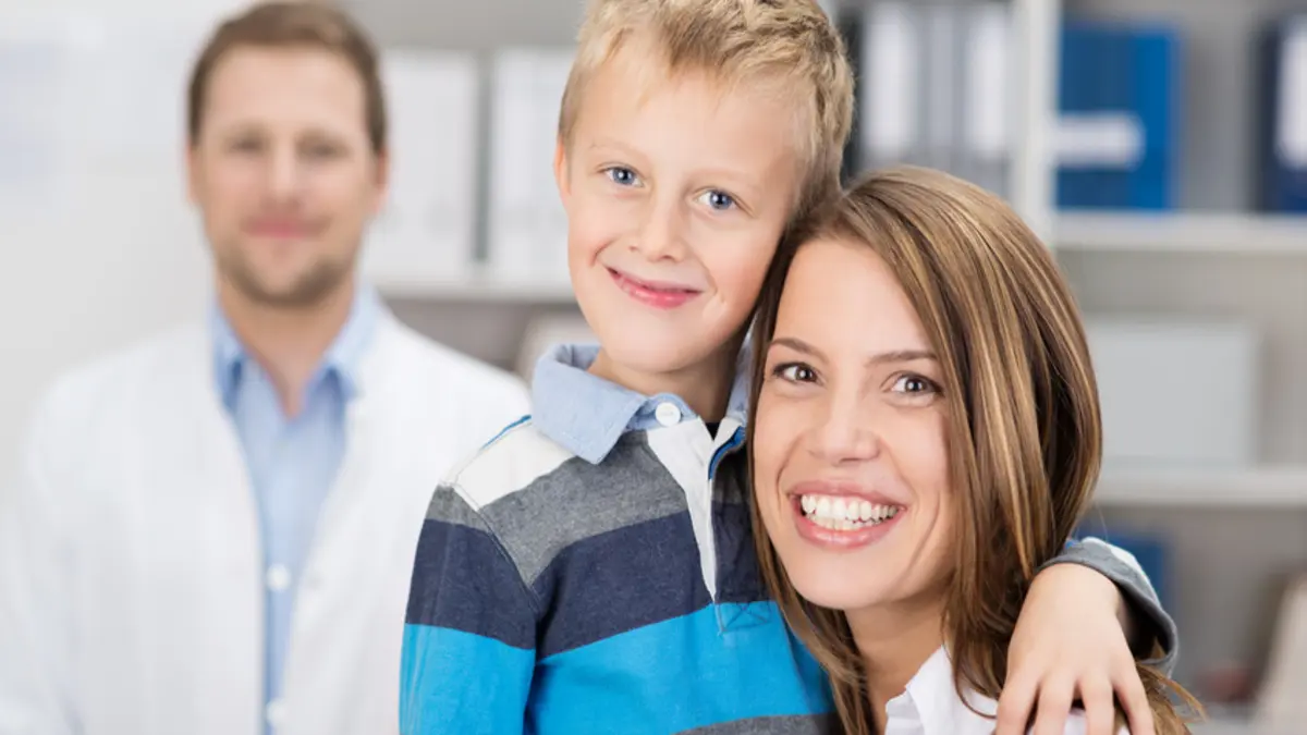 Mutter und Sohn in der Zahnarztpraxis. Sie lächeln in die Kamera, im Hintergrund der Kieferorthopäde