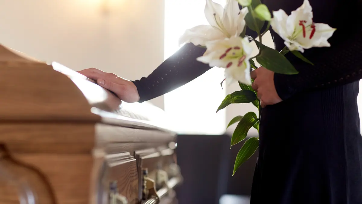 Eine Frau steht mit Blumen in der Hand an einem Sarg.