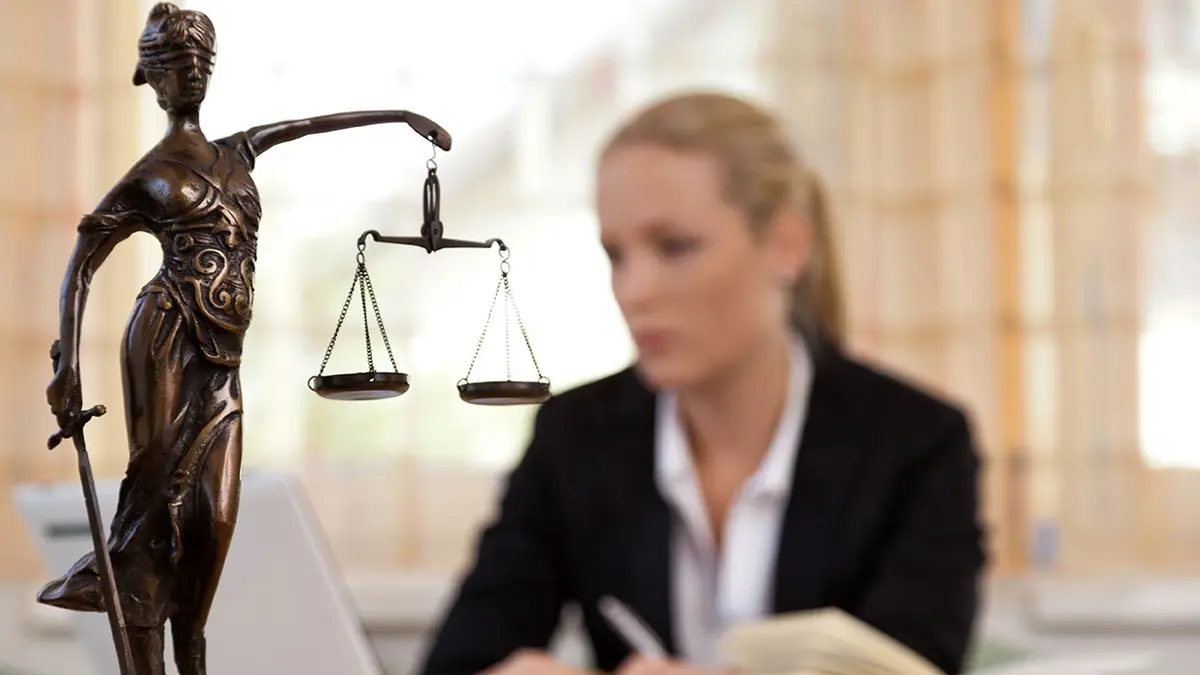 Justitia-Figur auf einem Tisch, im Hintergrund sitzt eine blonde Frau mit schwarzem Blazer