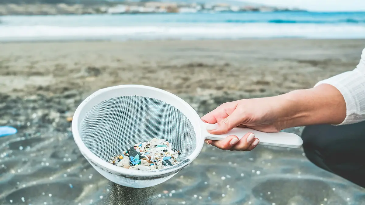 Frau am Strand sammelt mit einem Sieb kleine Plastikteilchen aus dem Sand