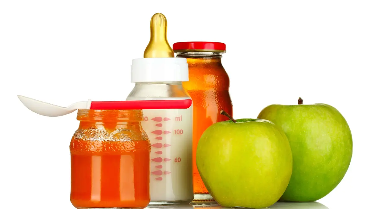 Im Vordergrund links steht ein Gläschen mit orange-rotem Babybrei. Darauf liegt ein Plastiklöffel für Babys. Rechts daneben liegen zwei grüne Äpfel. Im Hintergrund steht eine Babyflasche aus Glas, gefüllt mit Milch und eine kleine, verschlossene Glasflasche mit Apfelsaft.